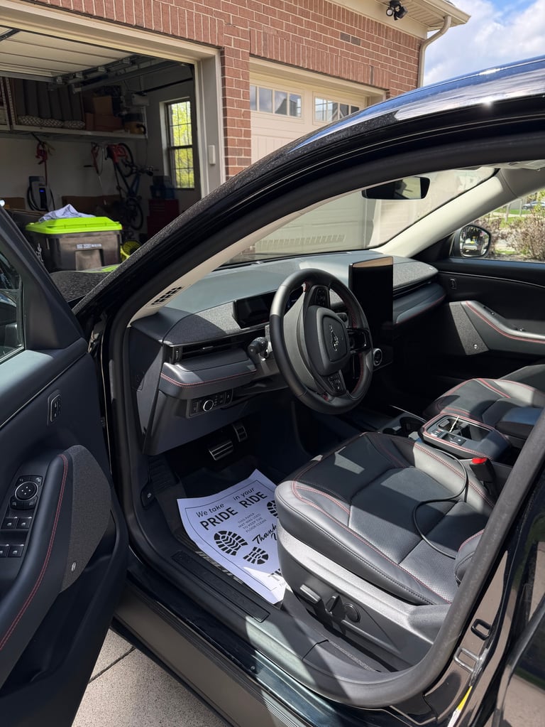 Open car door showing interior steering wheel and seats with a garage and brick house visible in the background