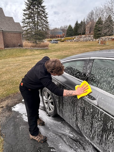 Person in black jacket washing a silver car with a yellow sponge in a residential driveway