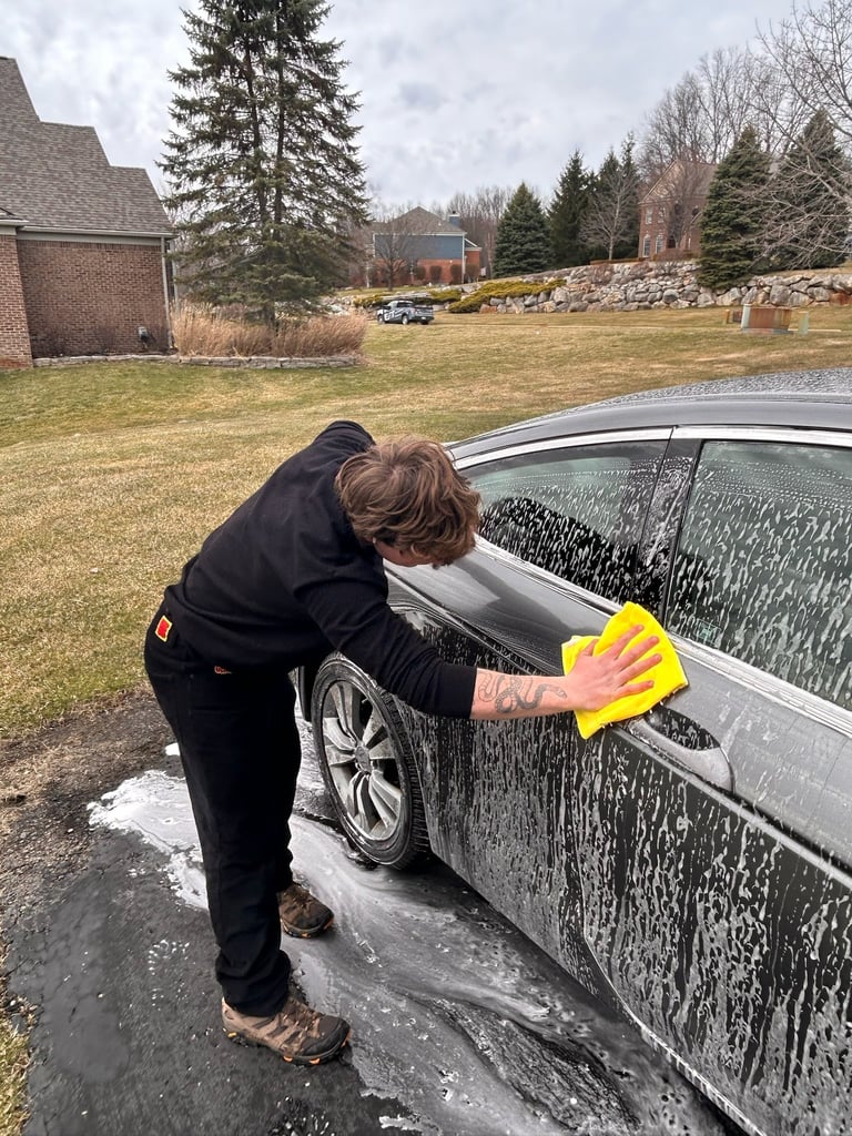 Person in black jacket washing a silver car with a yellow sponge in a residential driveway