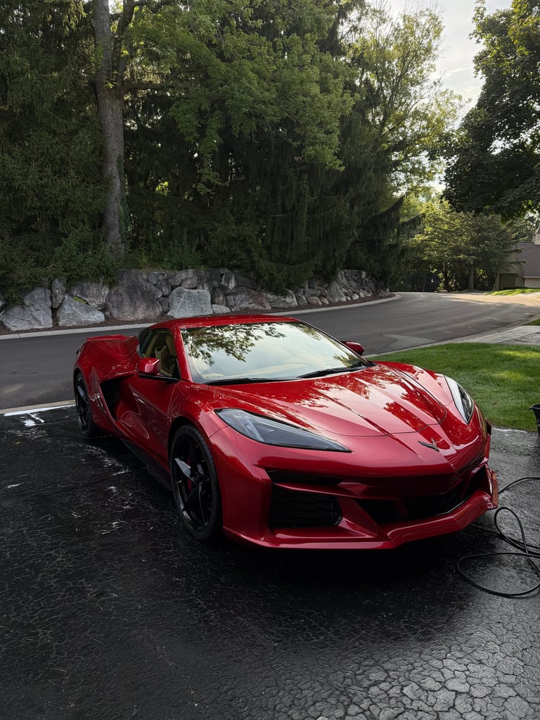 Red sports car parked on driveway with charging cable, surrounded by trees and landscaping