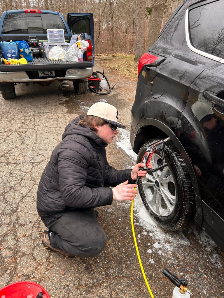 Person in black jacket and tan hat crouching to check tire pressure on black SUV with a measuring gauge in wooded area