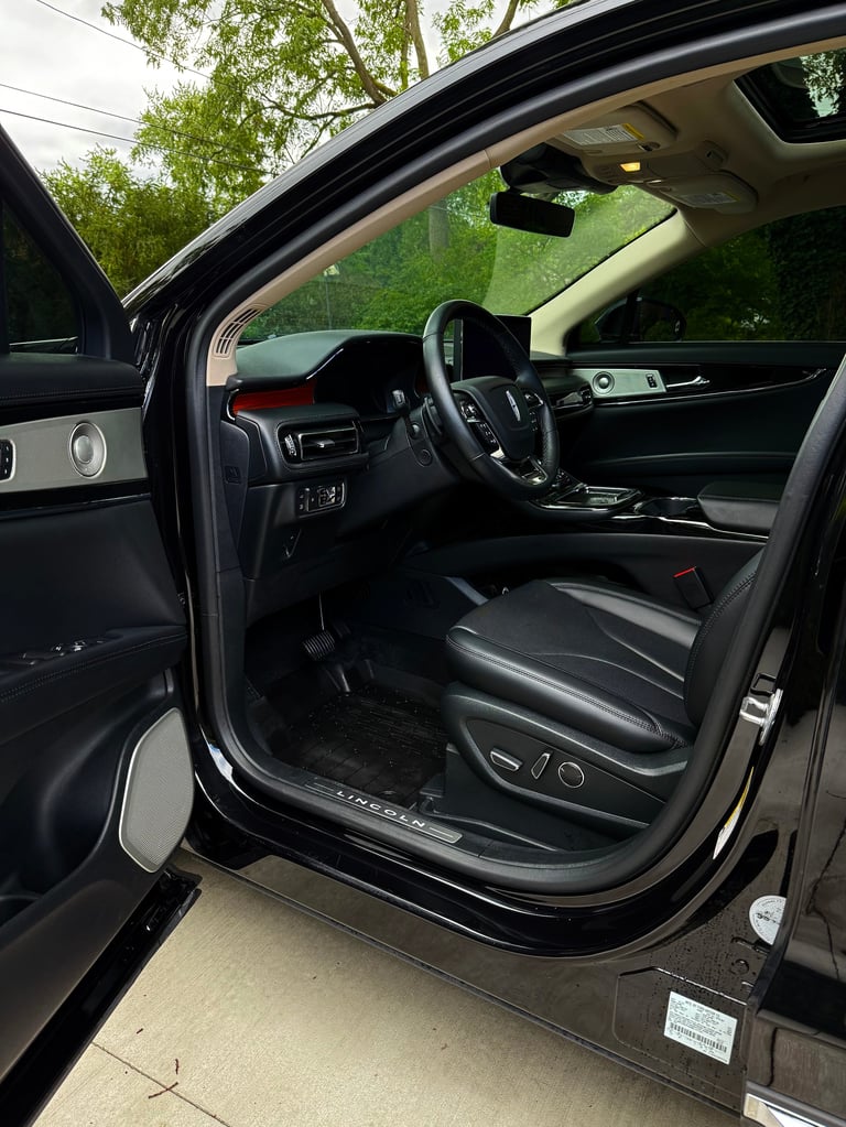 Open car door showing black leather interior with steering wheel, dashboard, and green trees visible in background