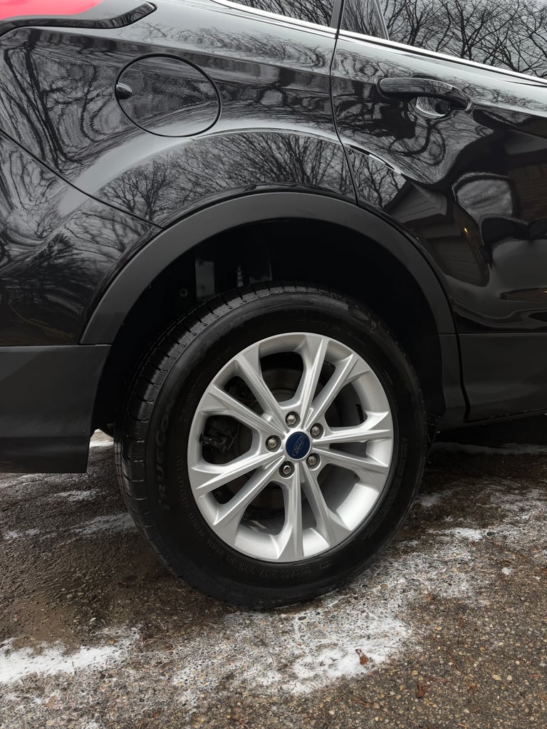 Close-up of a black SUVs silver alloy wheel and tire, with bare trees reflected in the glossy car body